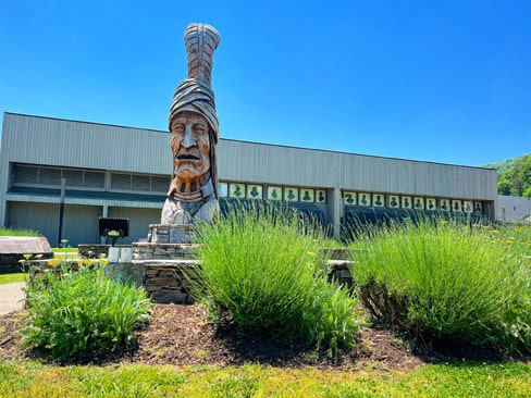 A large wooden statue of an indigenous man, wearing a tall, detailed headdress, stands in front of the Museum of the Cherokee People with circular decorative plaques. Green bushes embellish the foreground under