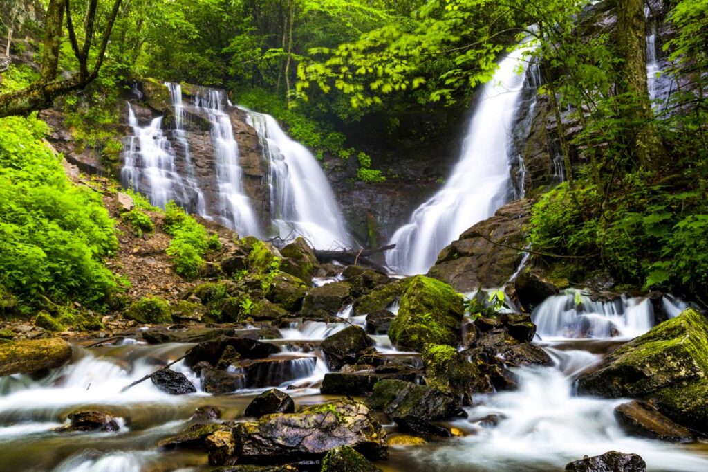 A lush, green forest setting with a multi-tiered waterfall cascading over rocks, surrounded by mist and vibrant foliage, with a stream flowing over jagged stones in the foreground at Soco Falls.