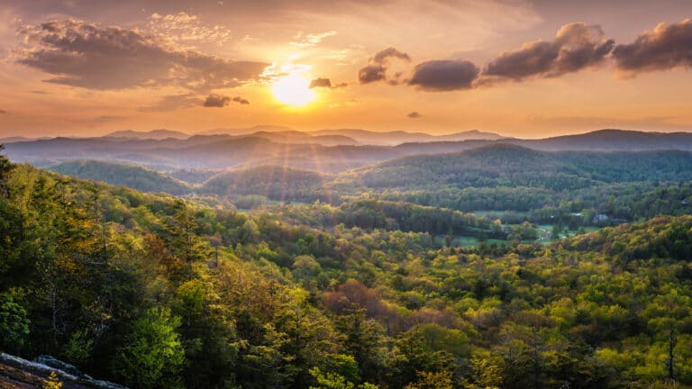 A scenic sunset over the lush, rolling Smoky Mountains with a rich carpet of green trees under a vibrant, orange-yellow sky filled with soft clouds.