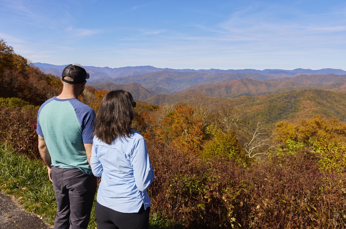 A man and a woman, tourists in Cherokee, stand looking out over vast, rolling hills covered in colorful autumn foliage under a clear blue sky.