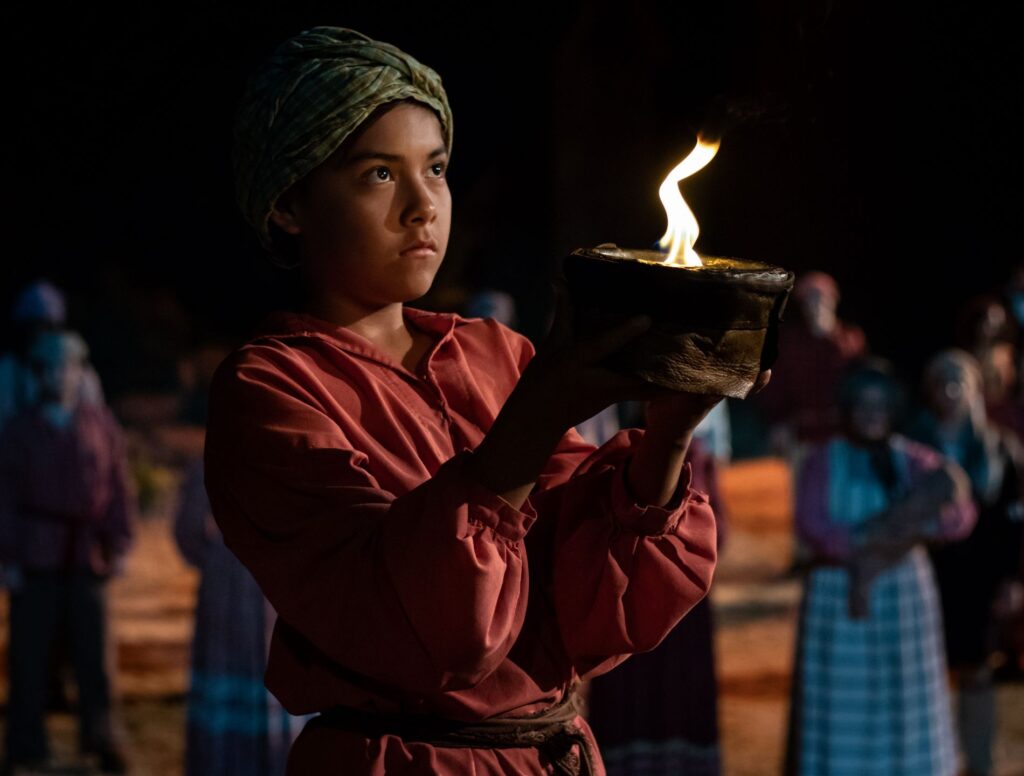 A young boy in traditional attire holding a flaming bowl during a nighttime performance of "Unto These Hills," surrounded by onlookers in various costumes.
