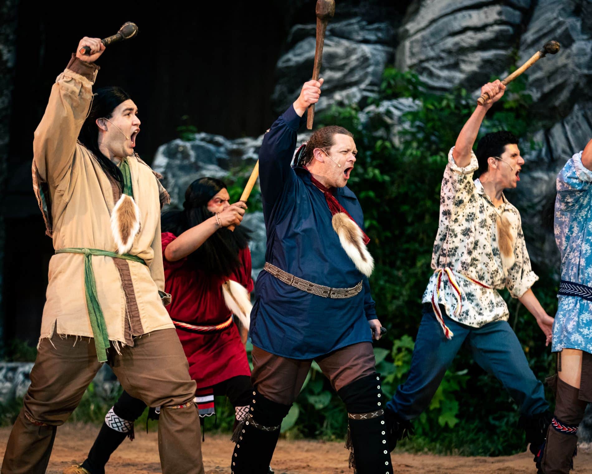 Four actors in costume perform on stage, energetically waving clubs as they act out a scene in the theatrical production of "Unto These Hills" with a rocky backdrop.