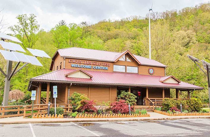 A wooden welcome center building in Cherokee, NC, with a gabled roof, surrounded by flowering shrubs and green hills, under a cloudy sky. A sign reads "Gateway to the Smokies Welcome