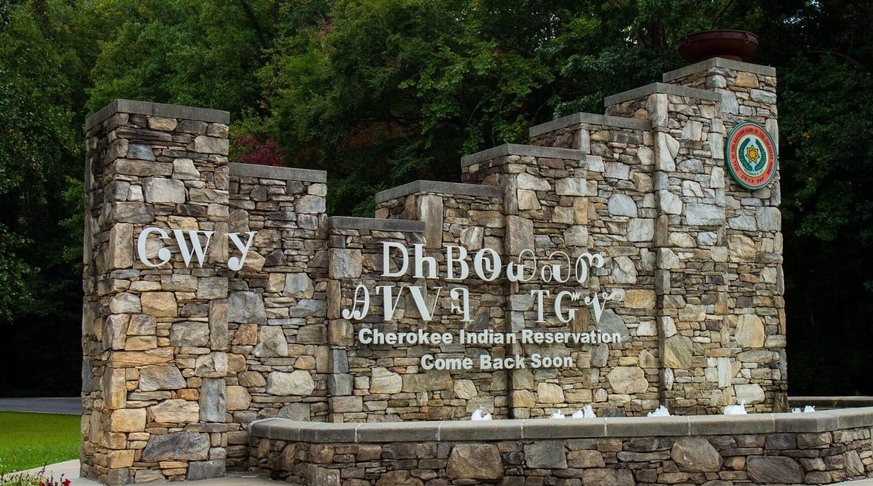 A stone entrance sign for the Cherokee NC Indian Reservation, with "Welcome" and "Come back soon" inscribed in English and Cherokee syllabary, adorned with the tribal seal. Green trees form