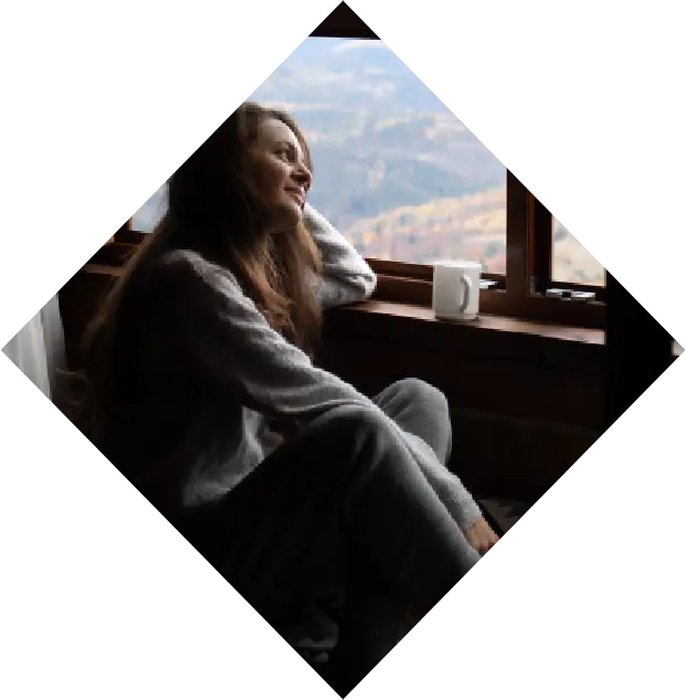 a woman in a cozy sweater sits by a triangular window in one of the cherokee cabins, gazing out at a mountainous landscape, with a mug beside her.